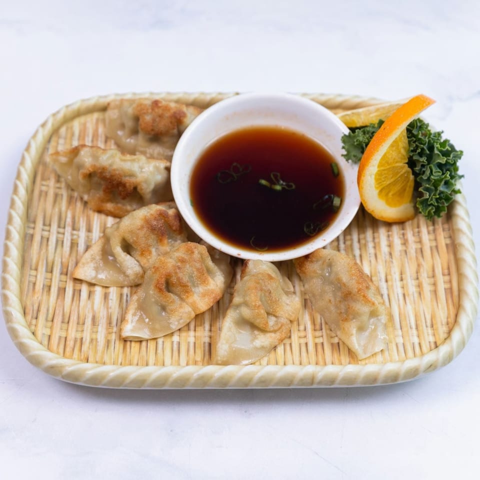 Pan-fried gyoza dumplings with a side of soy dipping sauce, garnished with orange and greens.