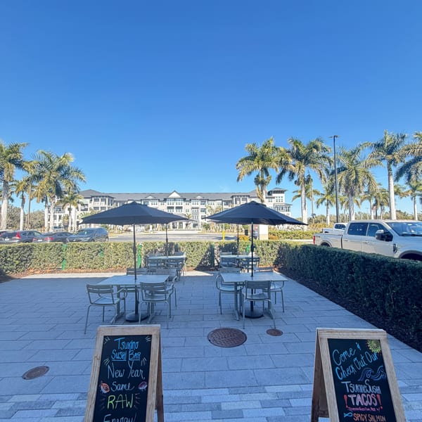 Tsukino Sushi Bar's outdoor patio with tables, umbrellas, and two chalkboard signs under a clear sky.