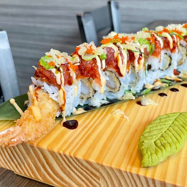 A long sushi roll with tempura shrimp, tuna, sauces, and roe, served on a wooden board.