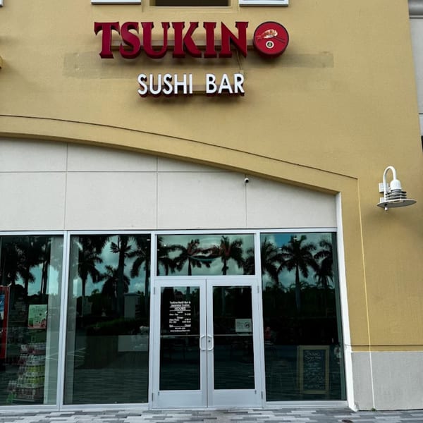 Exterior view of Tsukino Sushi Bar, featuring its red and white signage above glass doors reflecting palm trees.